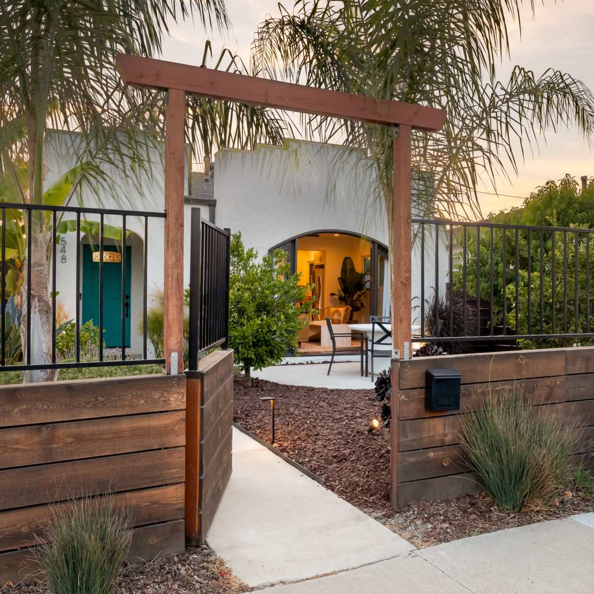 A wooden gate leading to a landscaped pathway that connects to a modern house surrounded by palm trees.