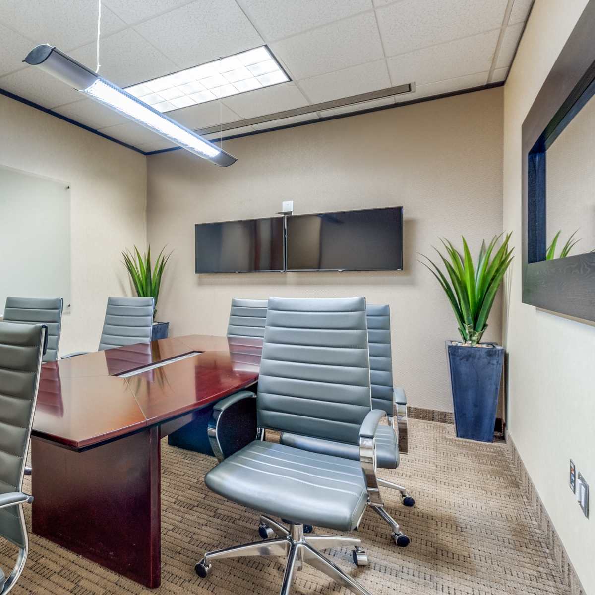 A modern conference room with a wooden table, six metal chairs, a whiteboard, and two wall-mounted screens.