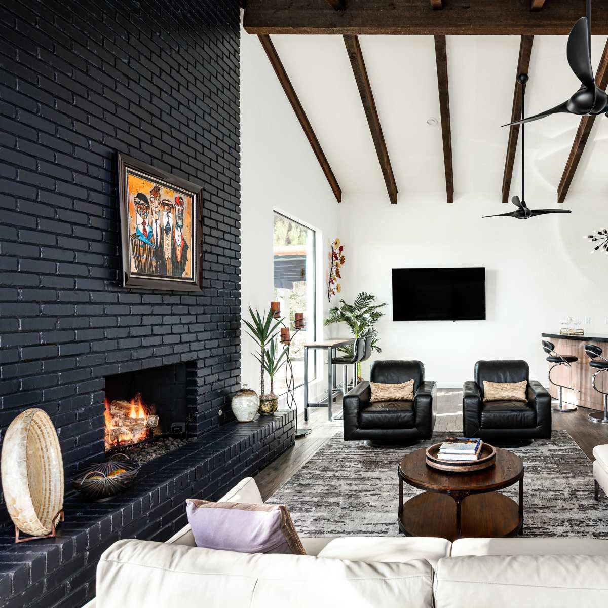 A modern living room featuring a white sectional sofa, a round coffee table, a black brick fireplace, and a flat-screen TV mounted on the wall, with exposed wooden beams overhead.