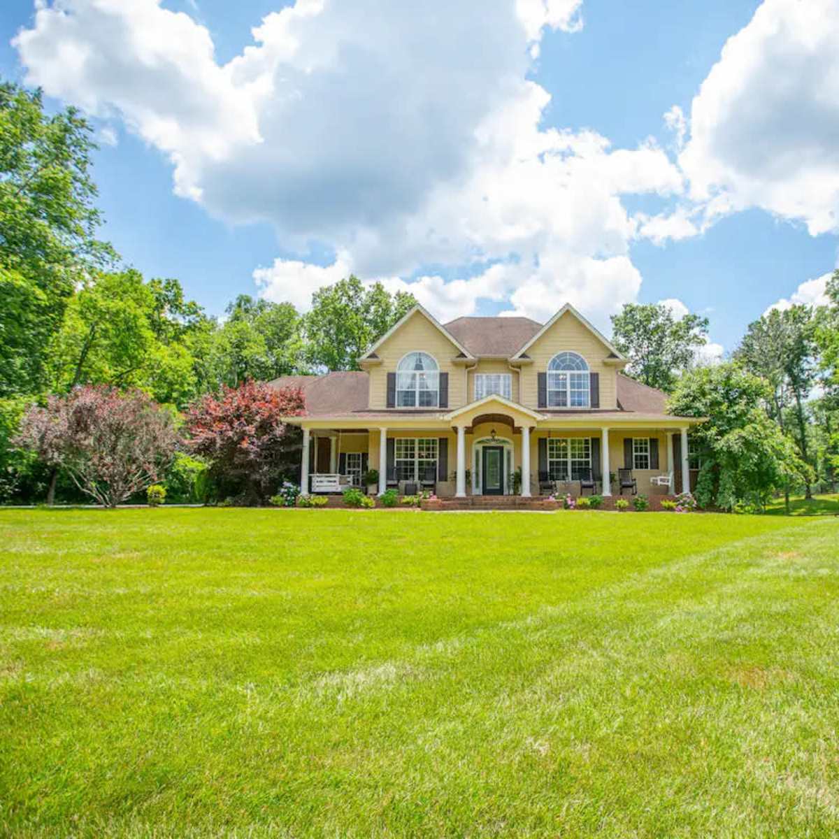 A two-story yellow house with a prominent front porch is situated on a large green lawn surrounded by trees.