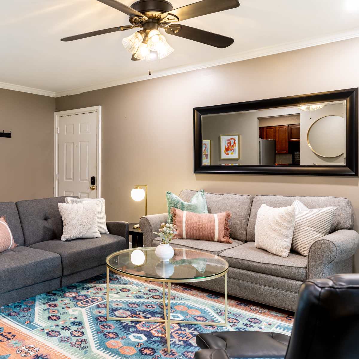The living room features a gray sectional sofa and a gray couch, with a glass coffee table centered on a colorful area rug, and a doorway leading to a bedroom in the background.