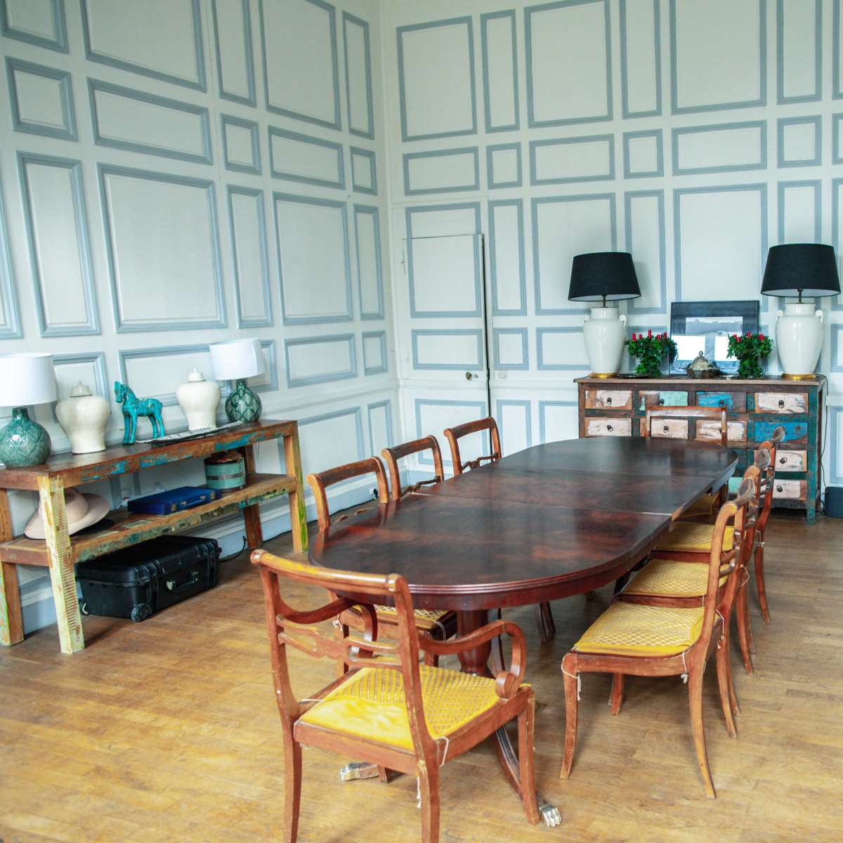 A spacious dining room features a large wooden table surrounded by eight chairs, with a sideboard and shelves displaying decorative items and plants.