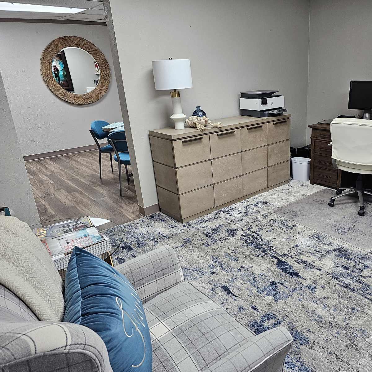 A small office space features a wooden desk with a computer, a printer, a light-colored sofa, a dresser, and a round mirror on the wall.