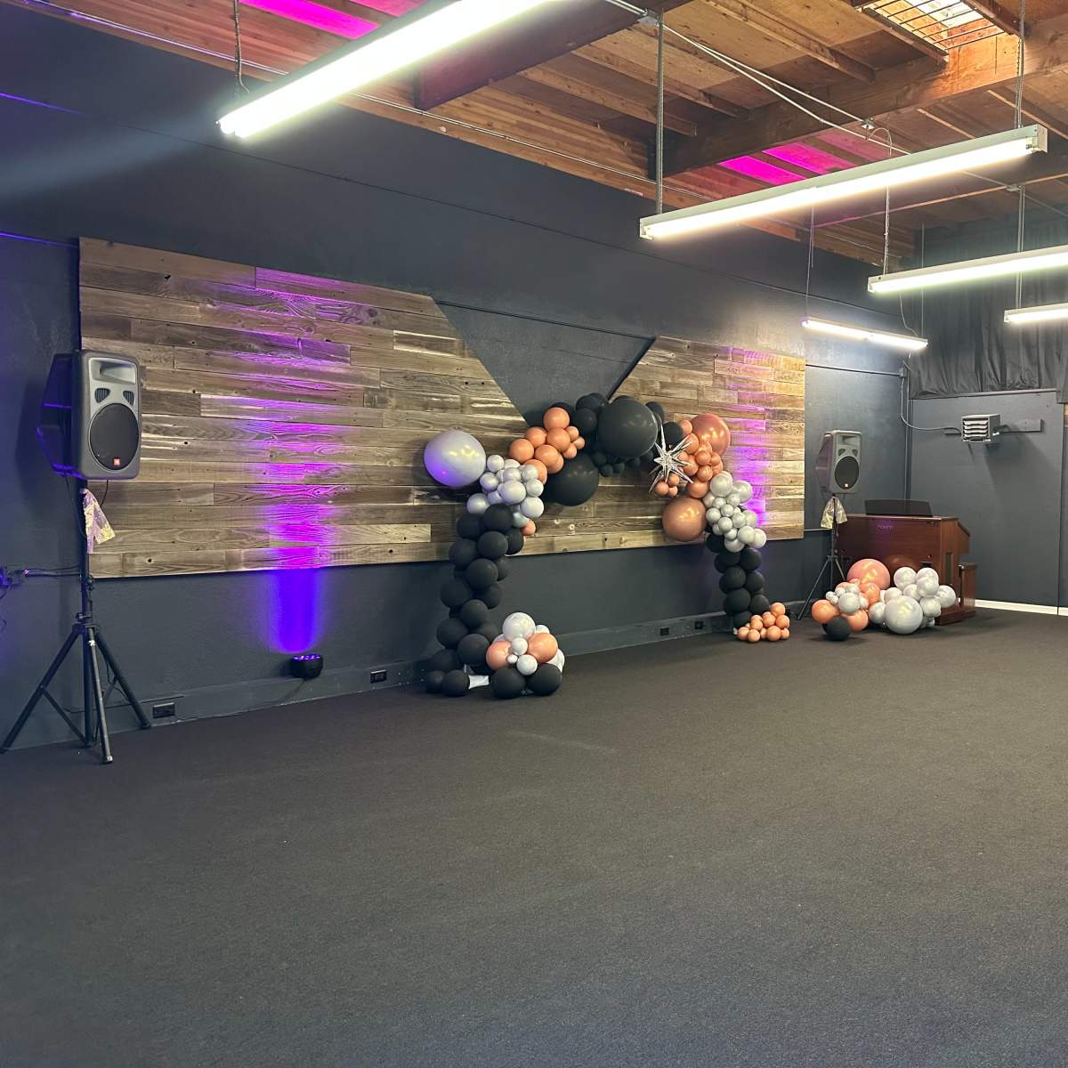 A spacious indoor venue with dark carpeting, wooden wall decor, and a large balloon arrangement to the side, along with speakers and a table set up in the background.