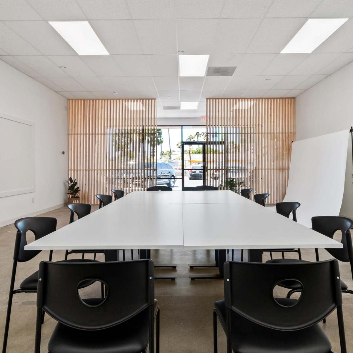 A modern conference room features a long white table surrounded by black chairs, with large windows allowing natural light to enter.