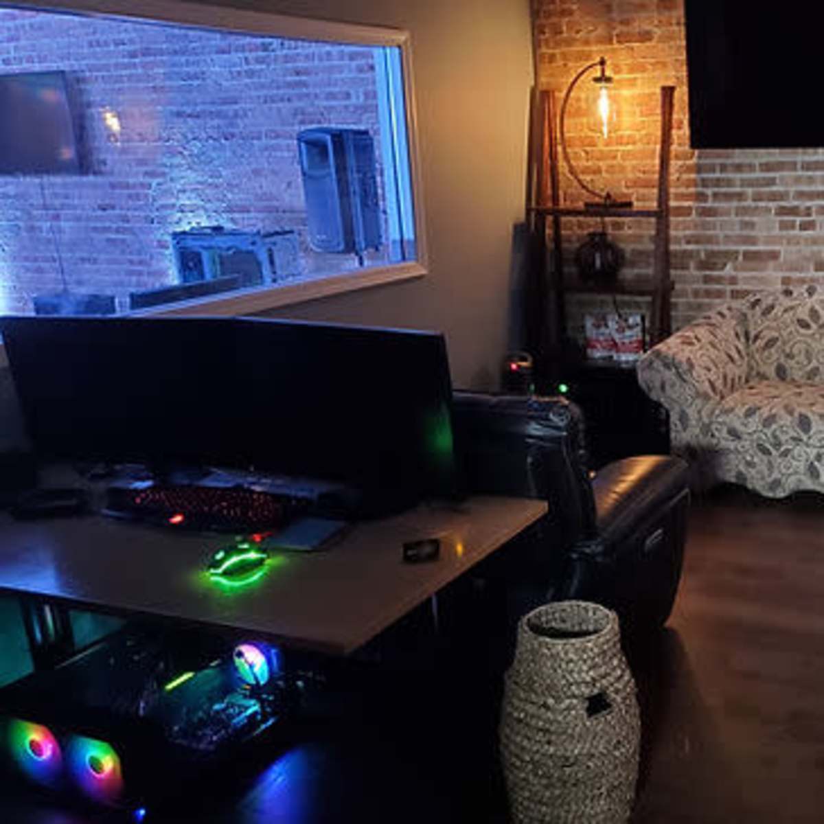 A cozy room with a sectional couch, a large computer setup with colorful lighting, and exposed brick walls.