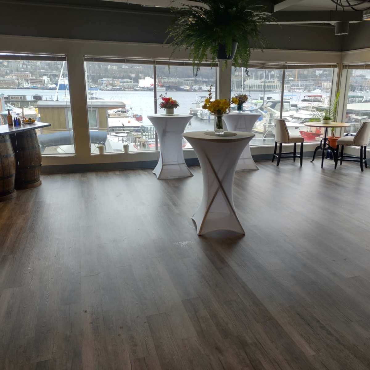 A spacious event room with several cocktail tables arranged around the perimeter, overlooking a marina through large windows.