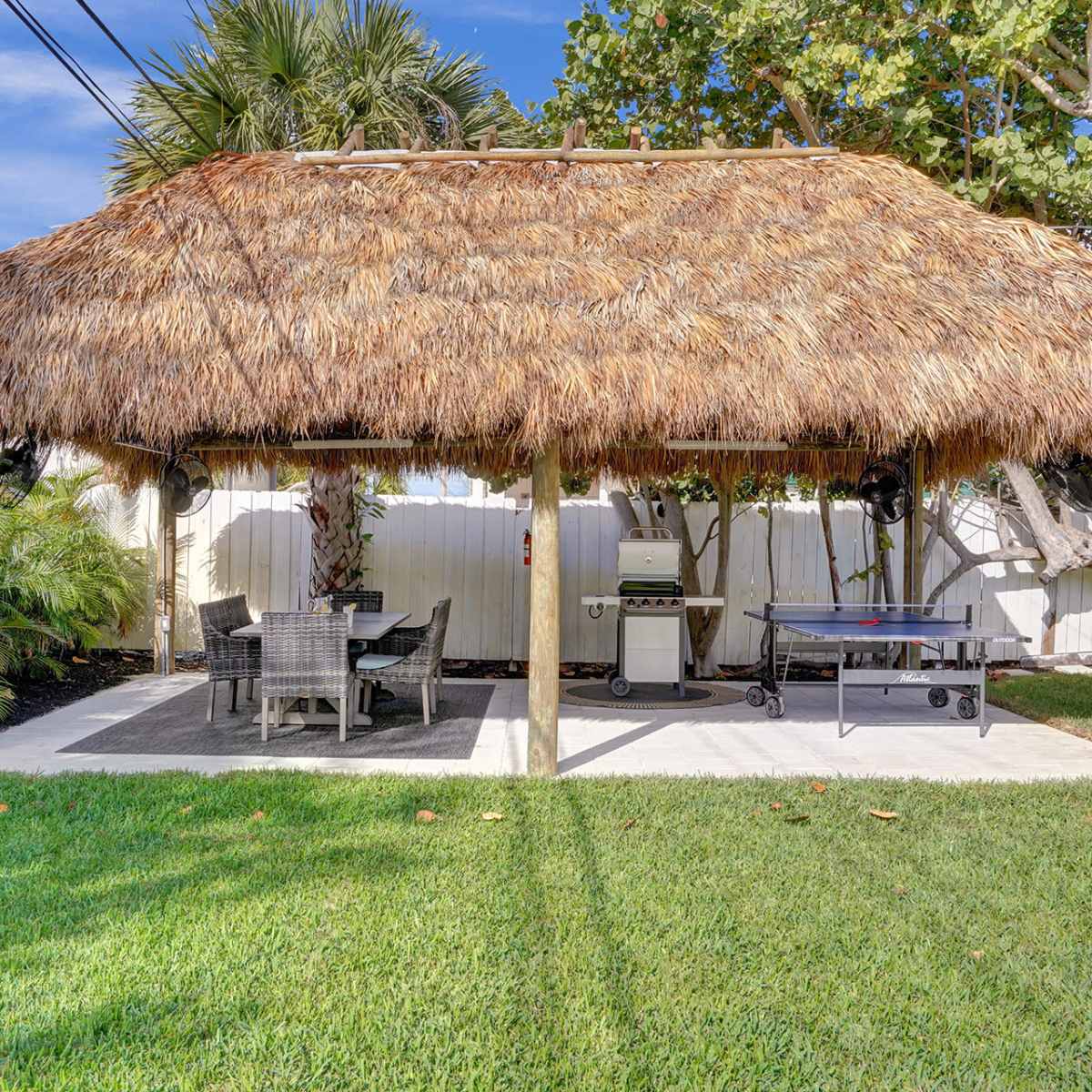 A thatched roof gazebo with a dining area and a grill is situated on a grassy lawn surrounded by palm trees.