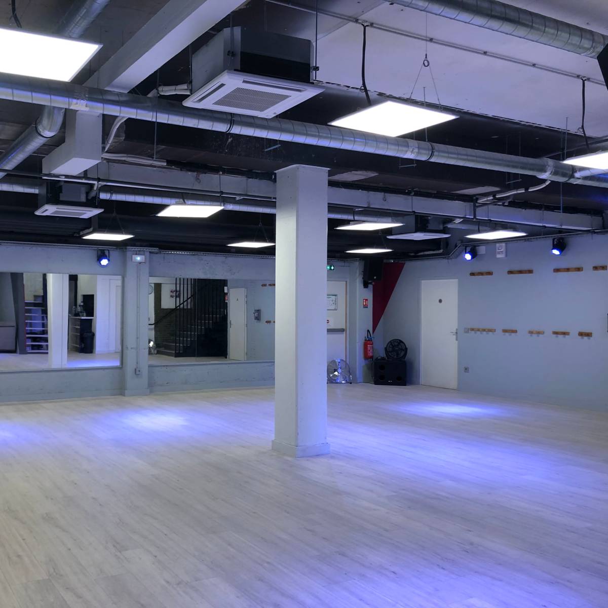 A spacious dance studio with wood flooring, bright ceiling lights, and large mirrors along one wall.