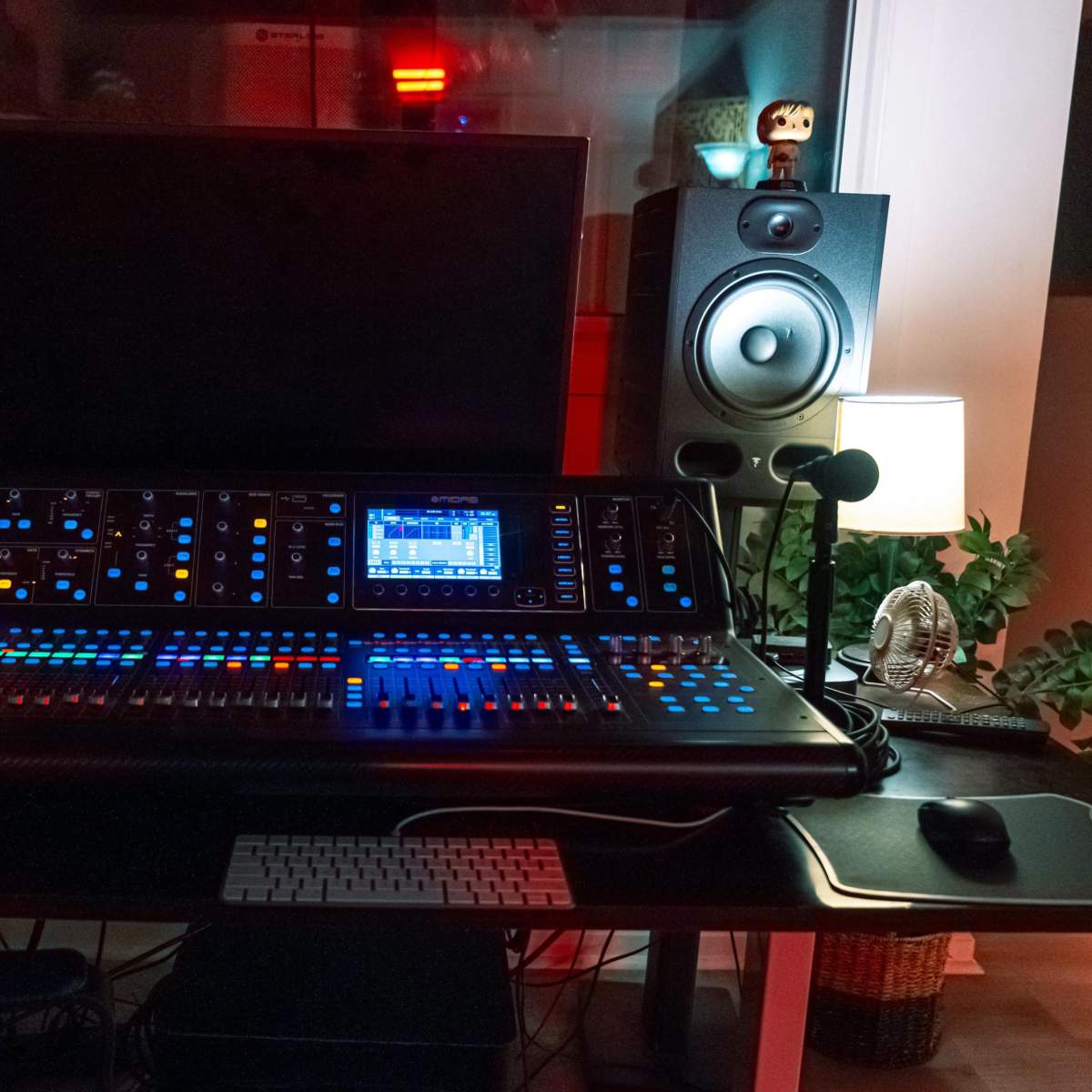 A music production workstation featuring a digital mixing console, studio monitors, a computer monitor, and a keyboard, set in a well-lit room with decorative items.