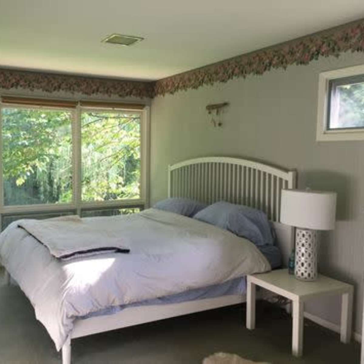 A bedroom features a bed with light blue bedding, a small bedside lamp, and large windows overlooking greenery.