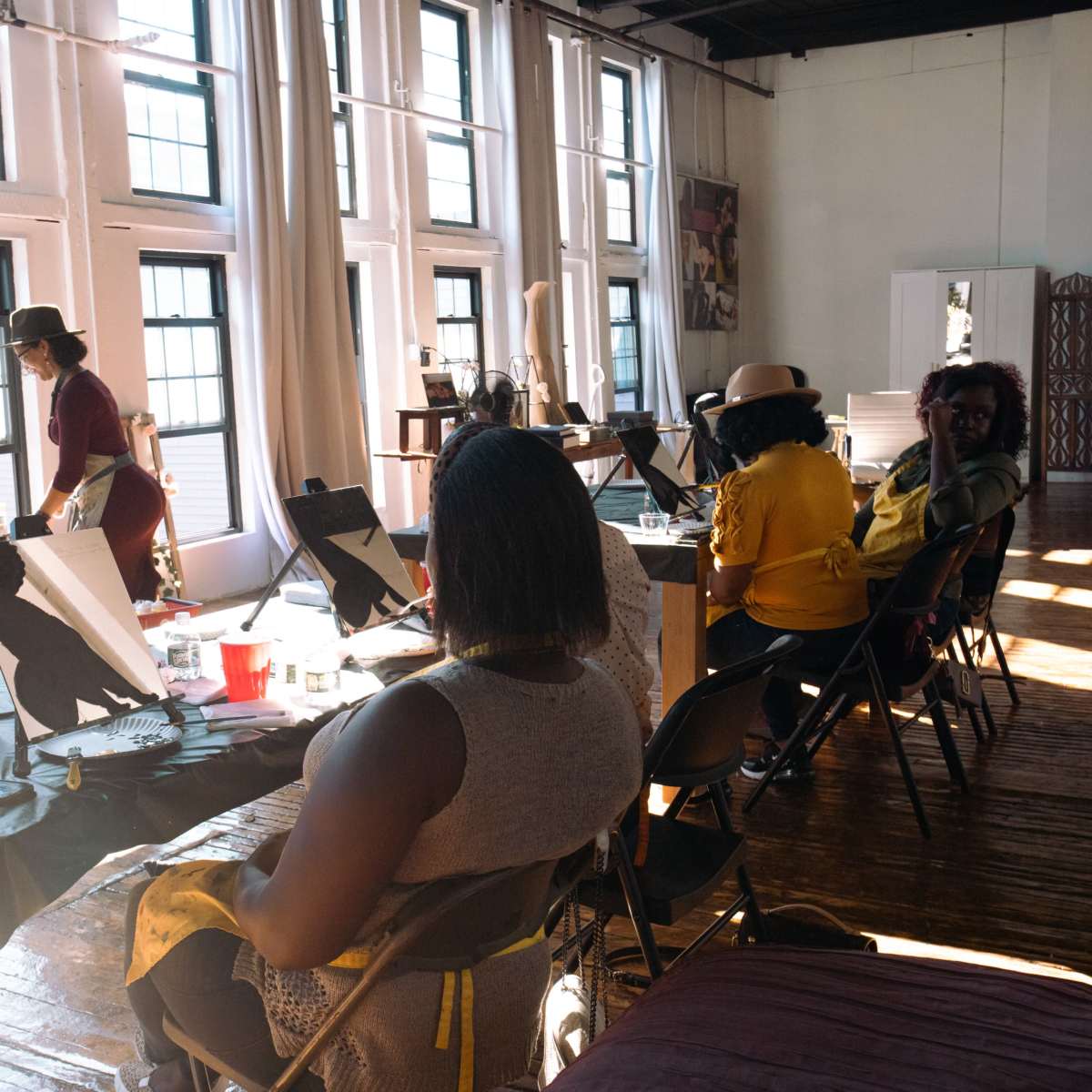 A group of individuals is engaged in an art class, seated at tables with easels in a sunlit studio featuring large windows.