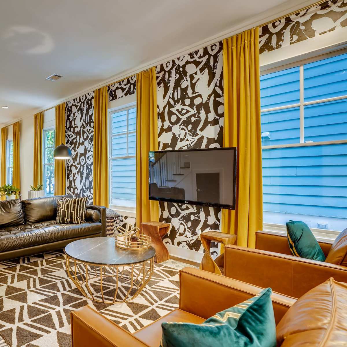 A modern living room featuring a leather sofa, two brown armchairs, a round coffee table, and a patterned accent wall, with large windows and yellow curtains.