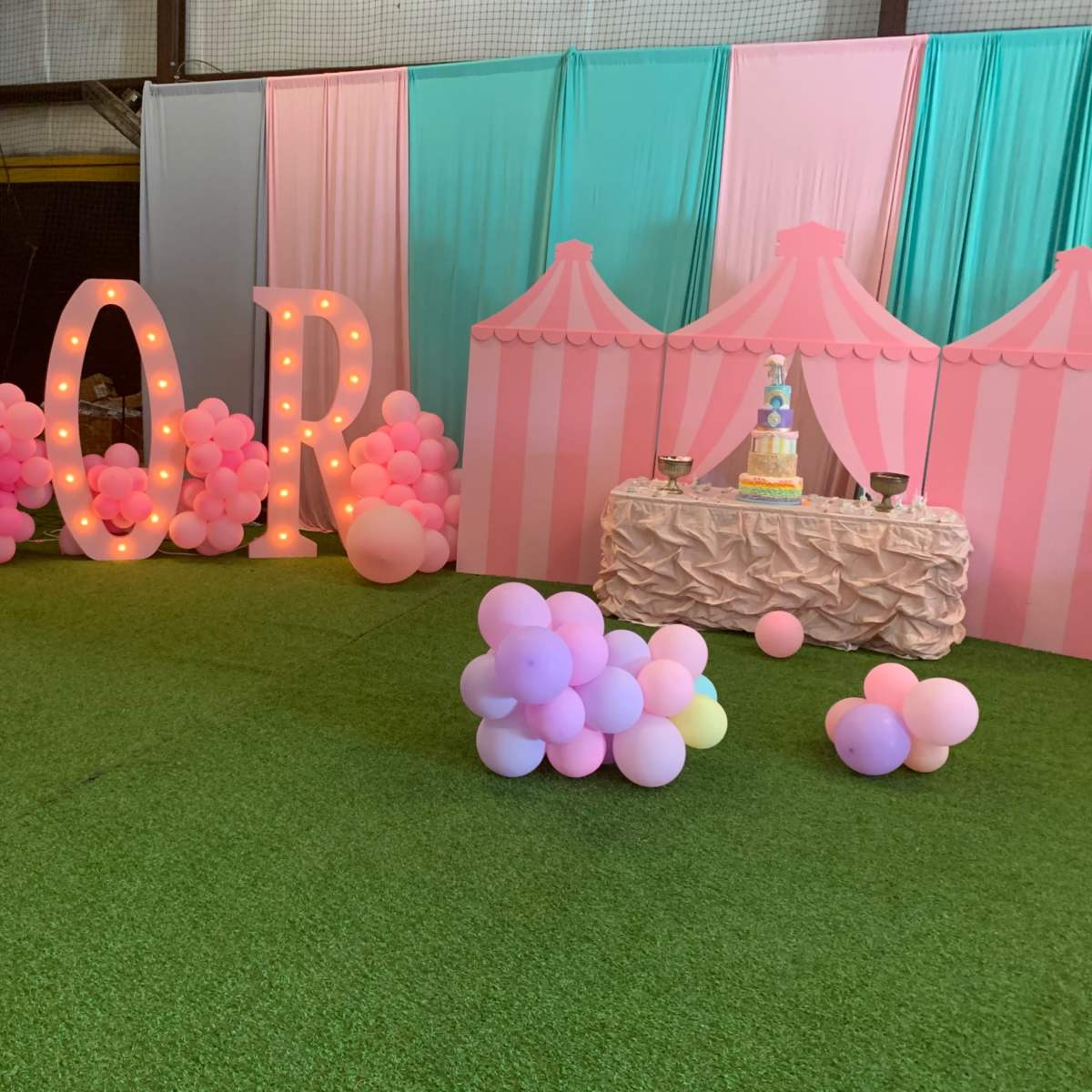The scene features large illuminated "DIOR" letters and a decorative display with pink and blue striped tents, complemented by a table showcasing a cake and colorful balloons on the ground.