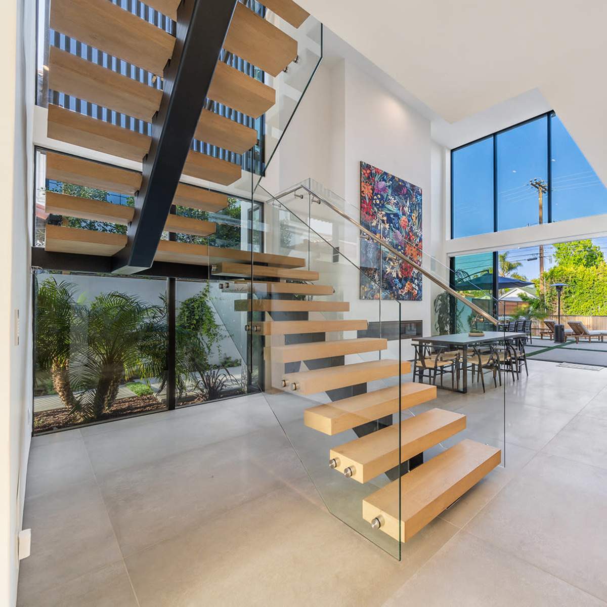 A modern interior space featuring a floating staircase with wooden steps, large windows providing natural light, and a dining area with a table and chairs.