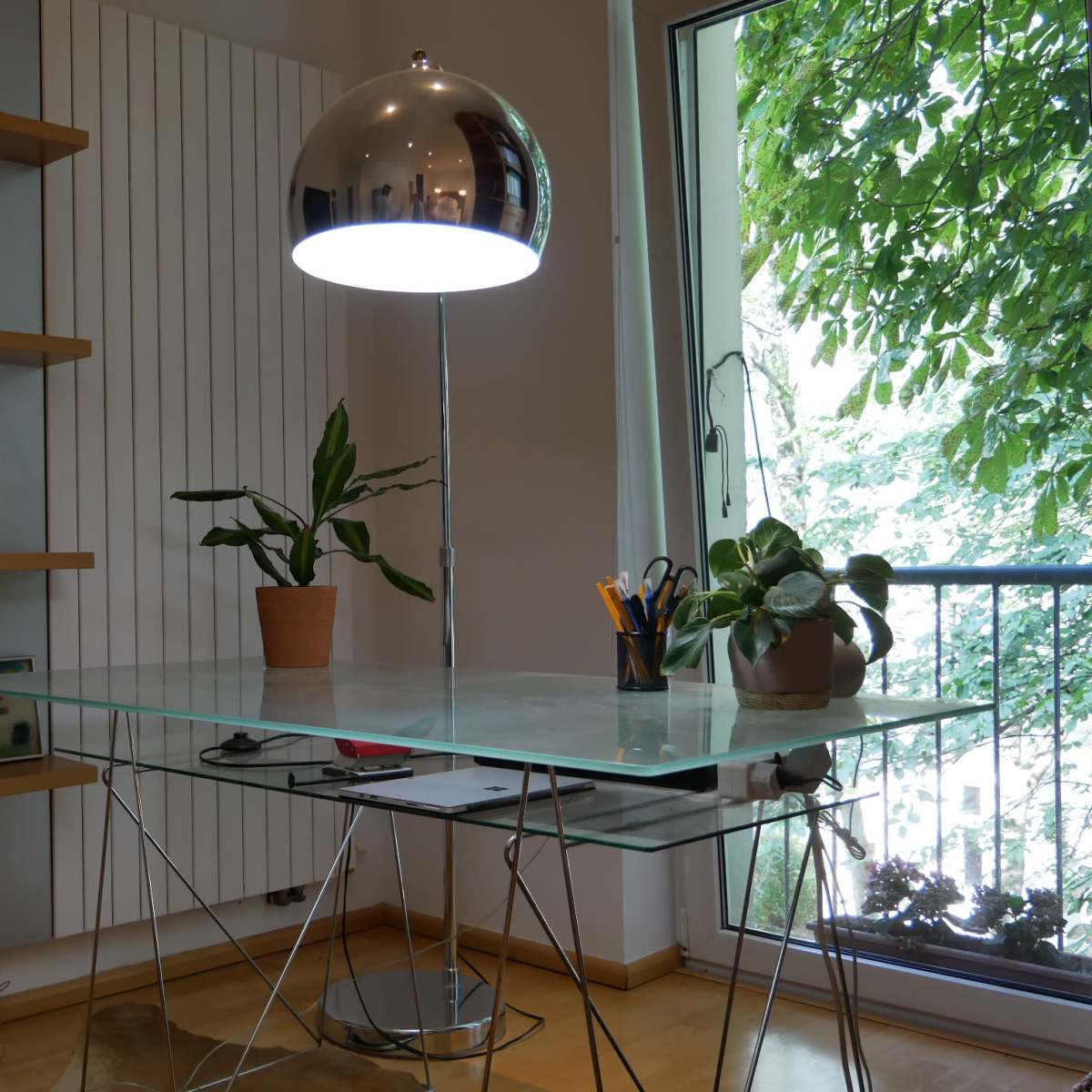 A modern glass desk is positioned next to a window with plants and a stylish lamp on top.