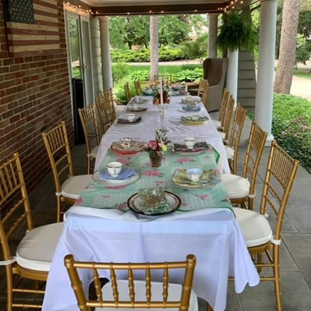 A long dining table is set with dishes and candles under a covered porch adorned with string lights.