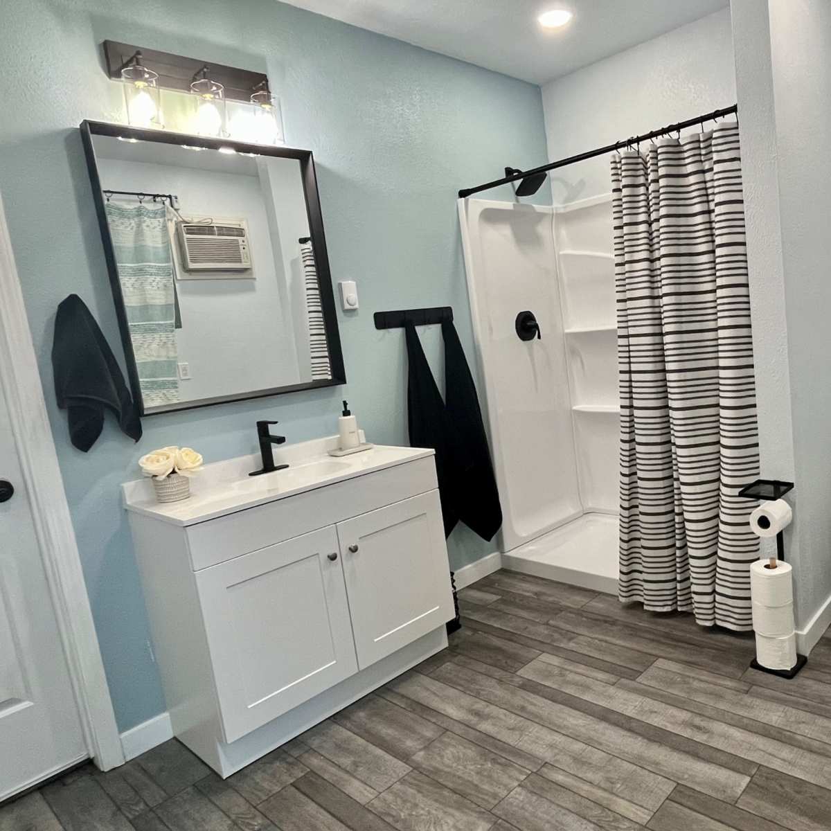 The bathroom features a shower with a striped curtain, a white vanity with a mirror, and a toilet, all set against light blue walls and wood-like flooring.