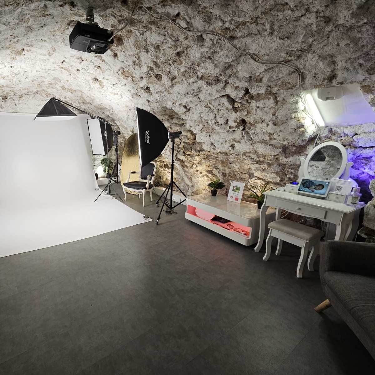 A photography studio set up in a stone-walled room, featuring softbox lights, a white backdrop, a vanity table, and minimalist furnishings.