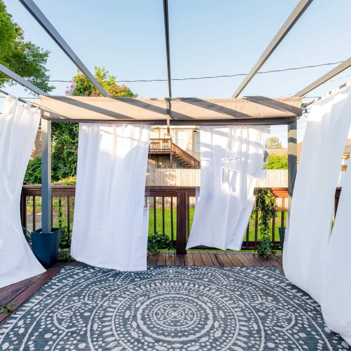 A wooden deck covered by a pergola with white curtains and a circular patterned rug on the floor.
