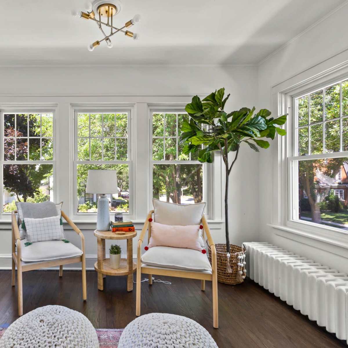 A bright sunroom features two armchairs, a small table with decor, a piano, and large windows overlooking green trees.