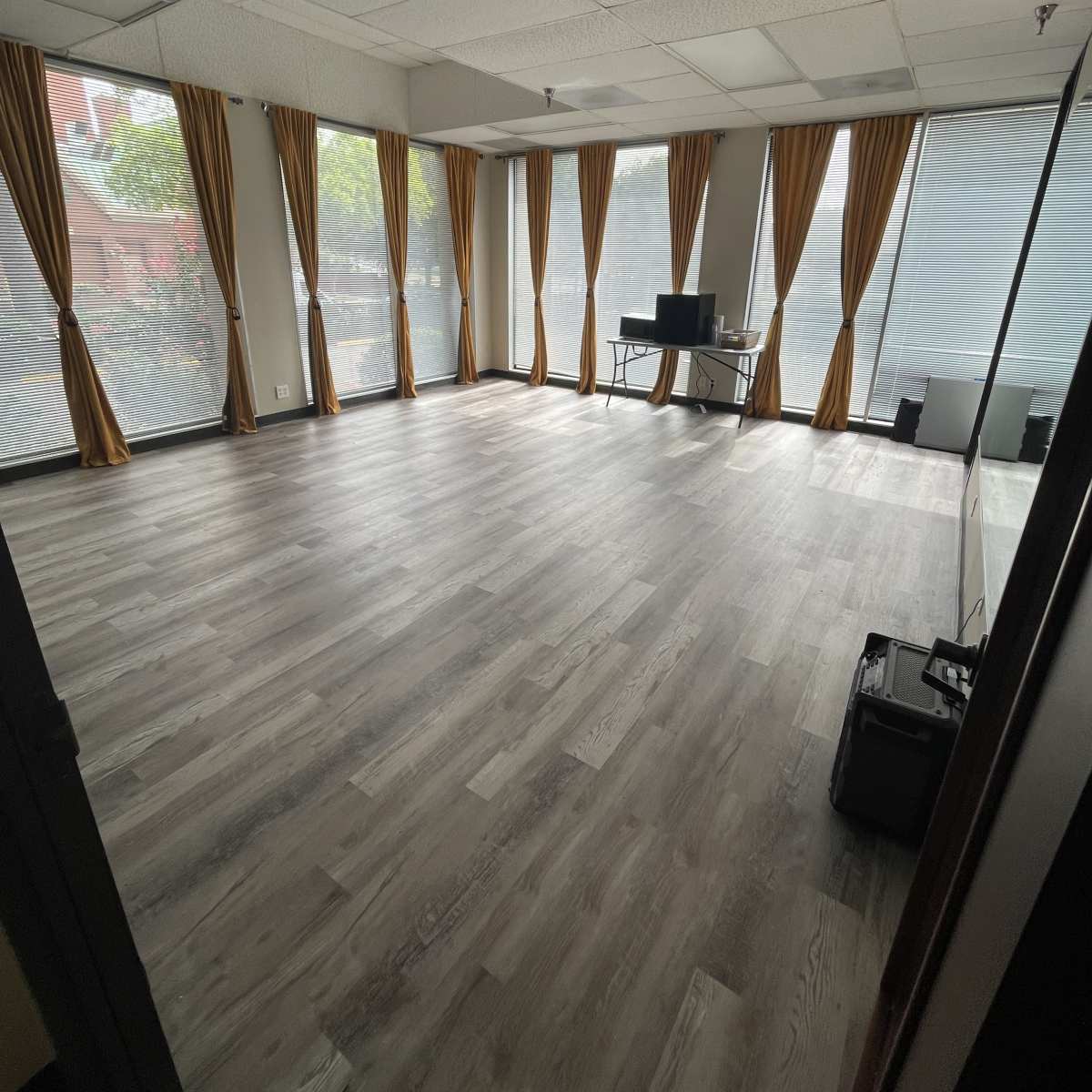 A vacant room with wooden flooring, large windows covered by sheer curtains, and a desk with a computer set up against one wall.