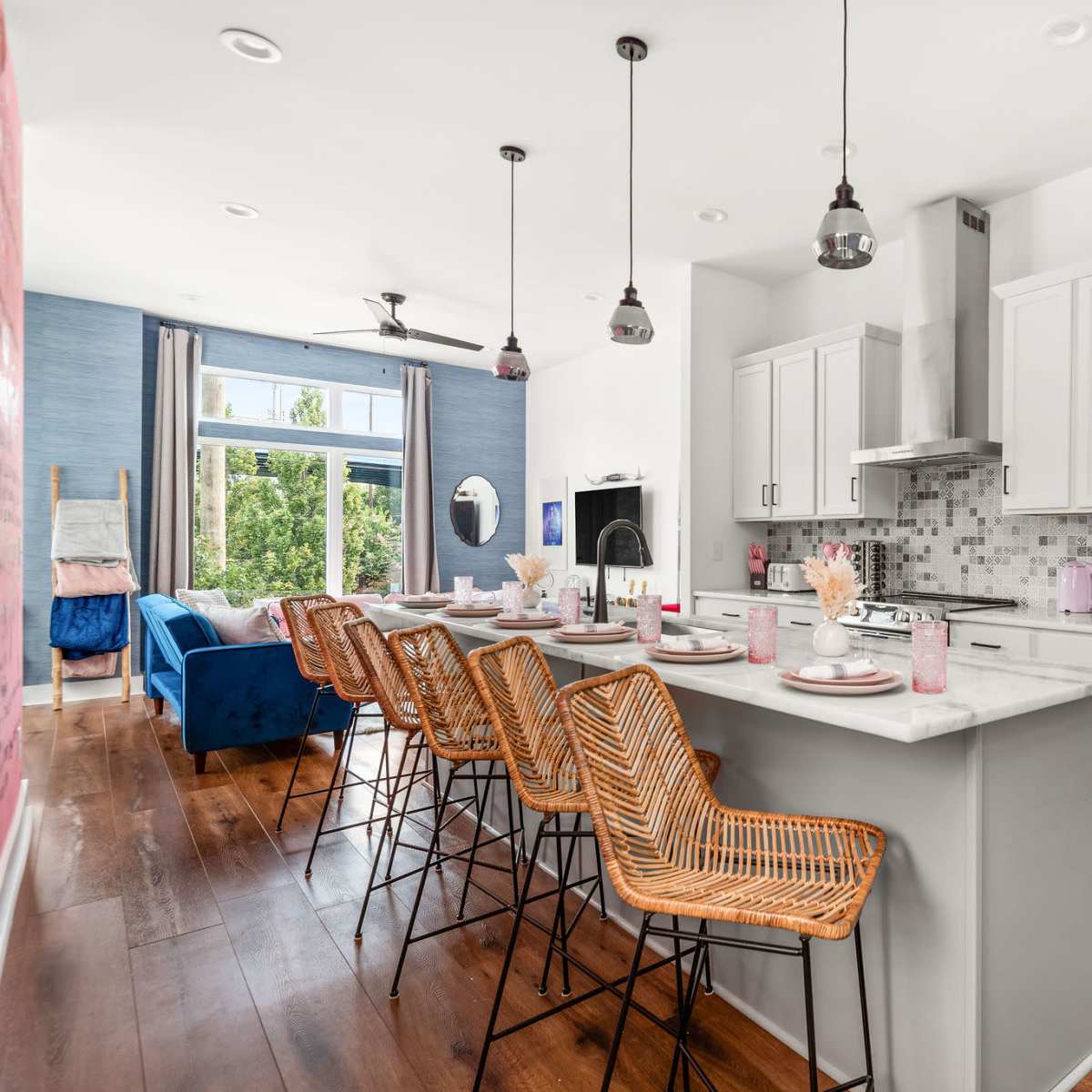 A modern kitchen with white cabinetry and a gray countertop features bar stools along a central island, with an adjacent living area visible in the background.