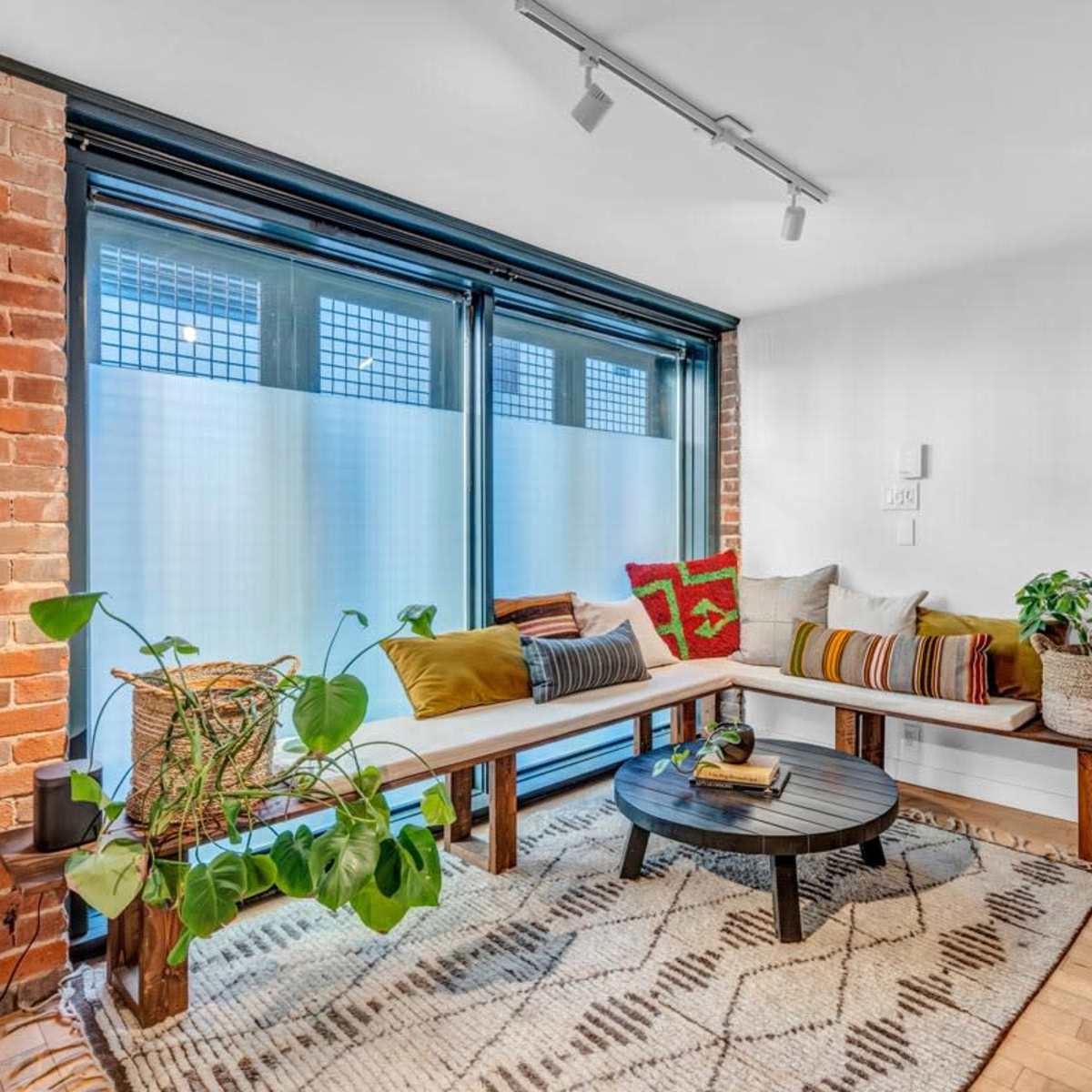 A cozy seating area features a built-in bench with cushions, a round coffee table, and plants, set against a backdrop of exposed brick walls and large windows.