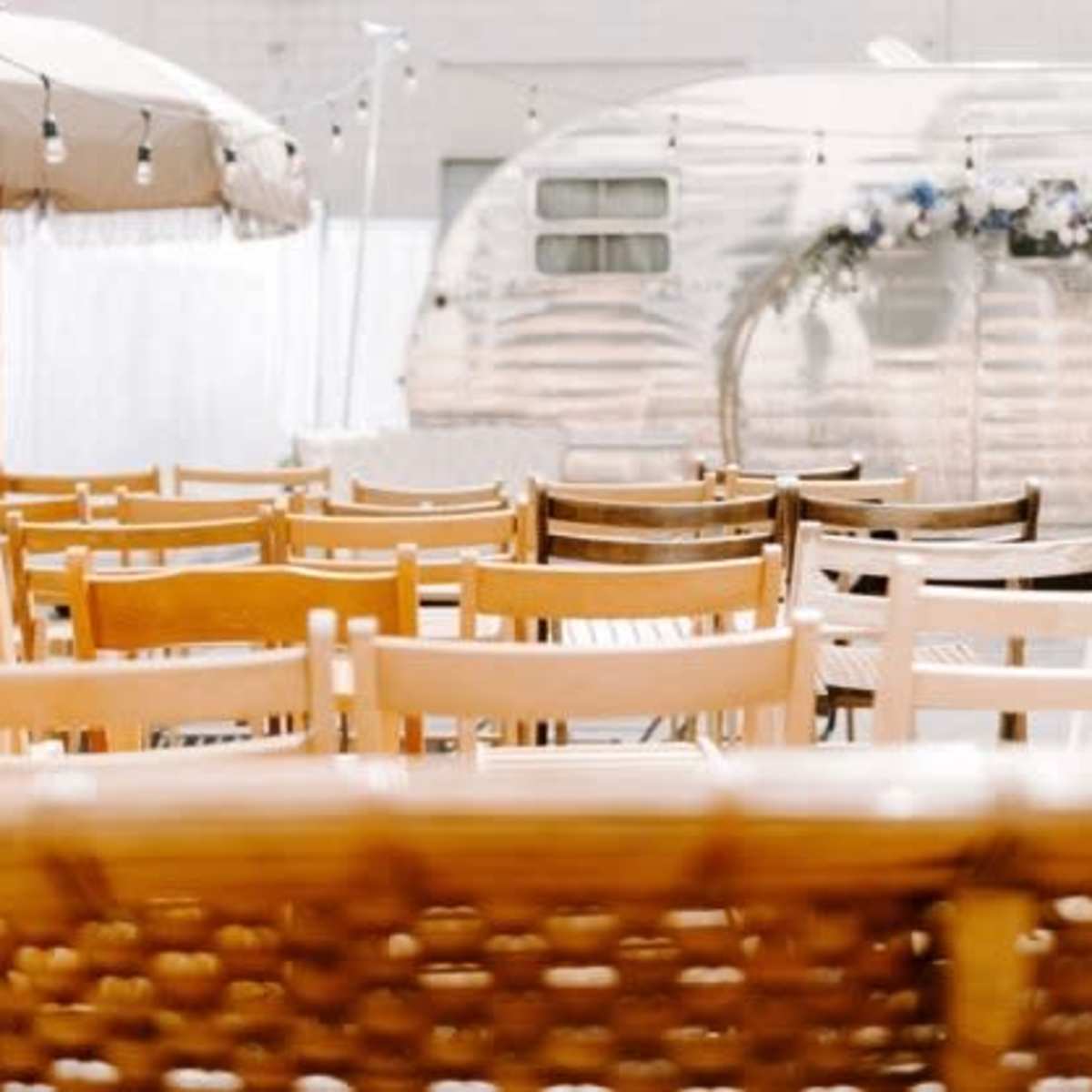 Rows of wooden chairs arranged for an outdoor event, with an Airstream trailer and a shaded area in the background.