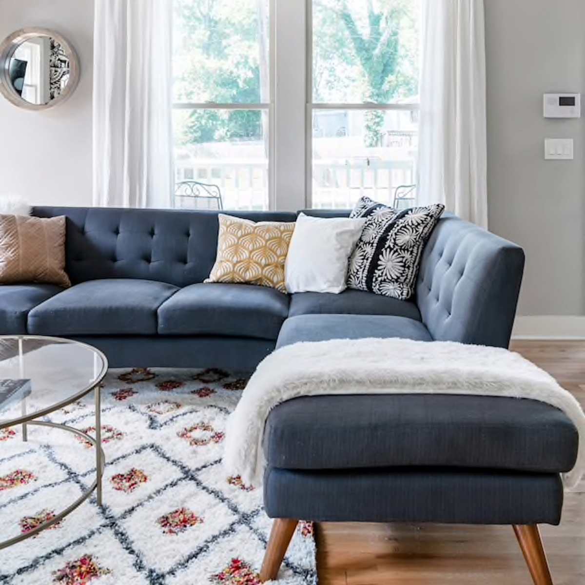 A modern living room with a sectional sofa, a round glass coffee table, and large windows dressed with white curtains.