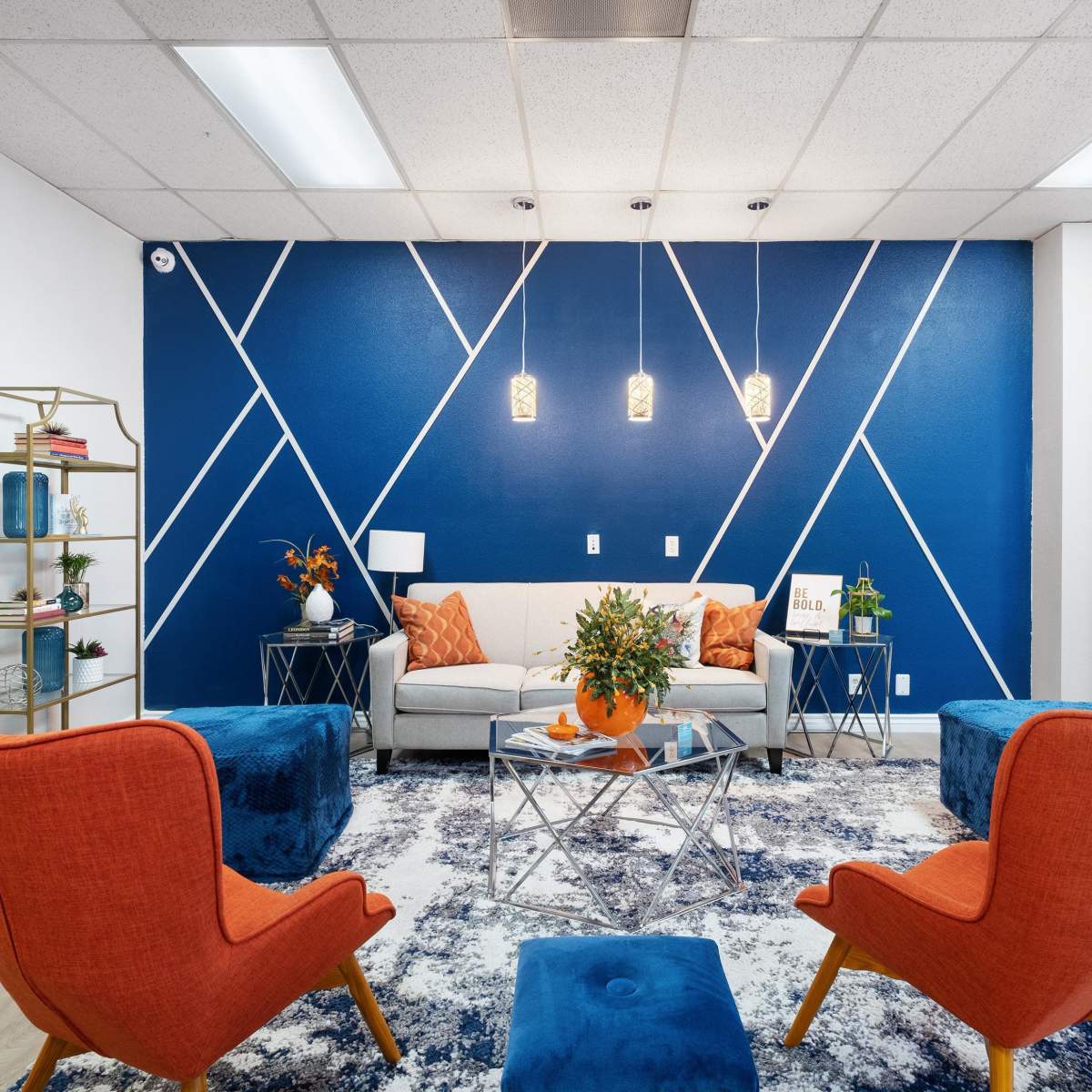 A modern seating area with orange chairs, a gray sofa, and a blue geometric accent wall, complemented by a decorative rug and plants.
