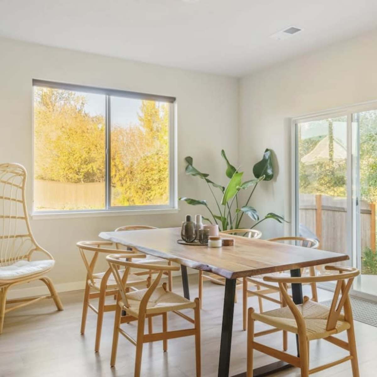 A dining area features a wooden table surrounded by chairs, with a large window and sliding door revealing a view of a backyard.