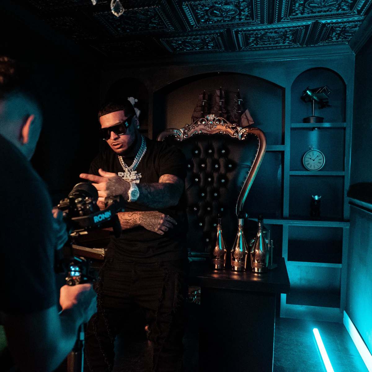A man in dark sunglasses poses confidently in front of a camera in a dimly lit, stylish room with a large, ornate chair and decorative bottles on a table.