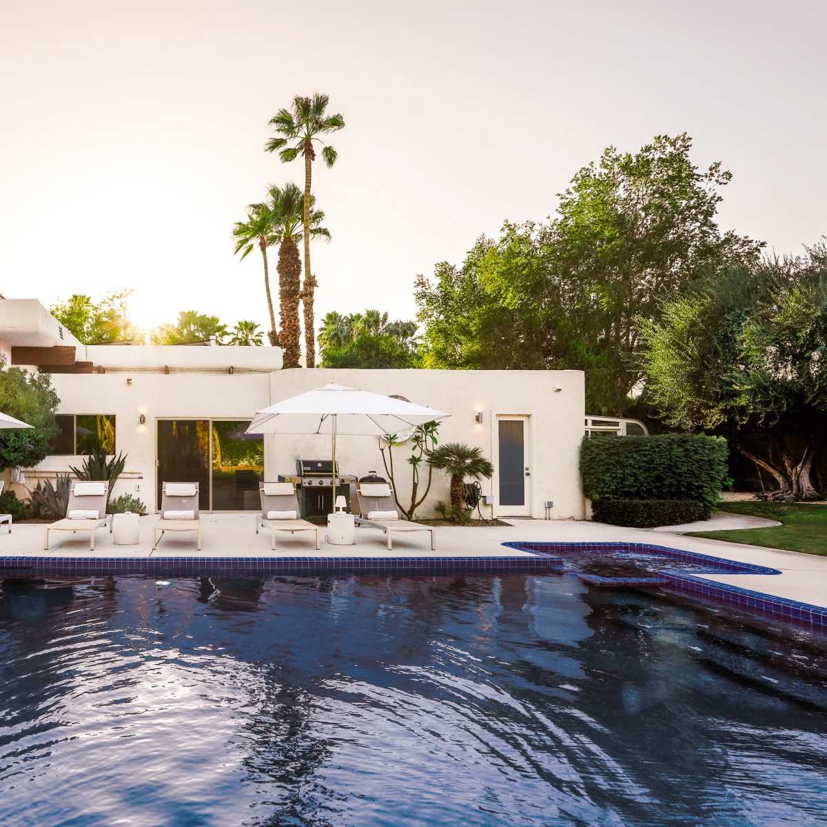 A modern home with a pool, lounge chairs, and umbrellas, set against a backdrop of palm trees and a bright sky.