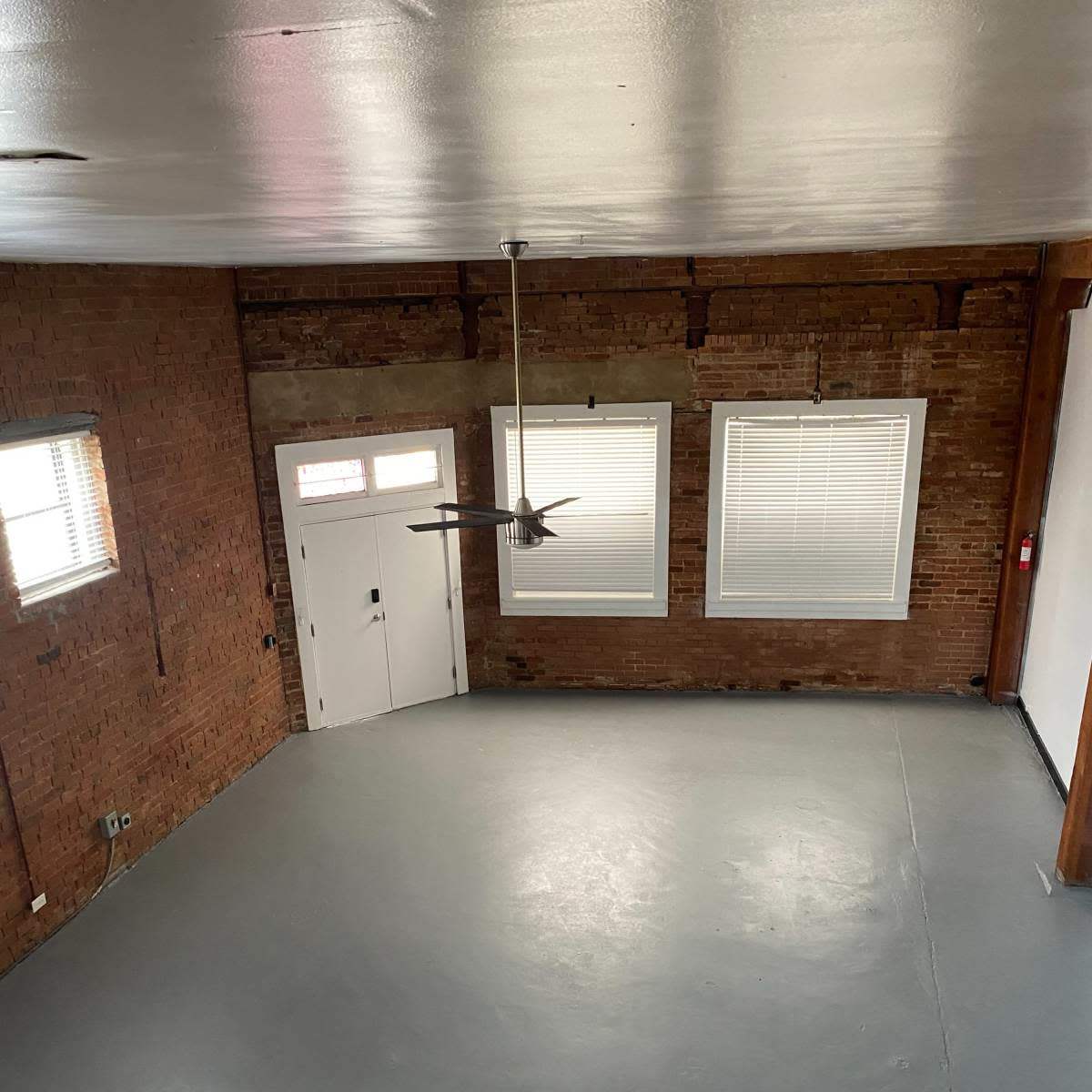 An empty room with exposed brick walls, a concrete floor, and two windows, along with a ceiling fan.