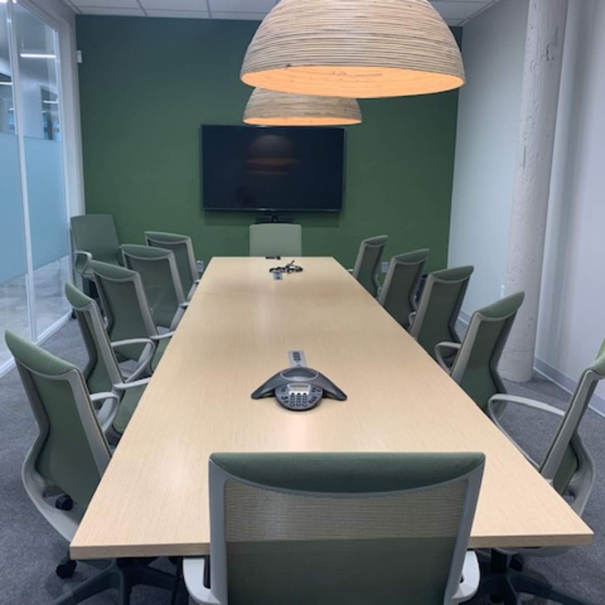 A large conference room features a long wooden table surrounded by green chairs, with two pendant lights hanging above and a wall-mounted screen at the front.