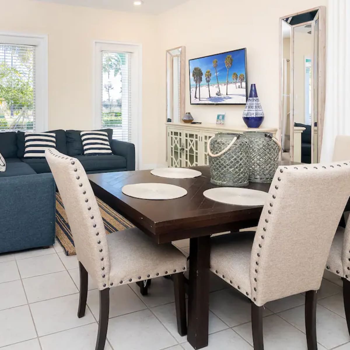 A living and dining area featuring a blue sofa, a dining table with beige chairs, and large windows providing natural light.