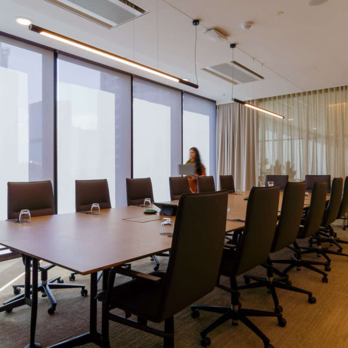 A modern conference room features a long wooden table surrounded by black chairs and large windows with sheer curtains.