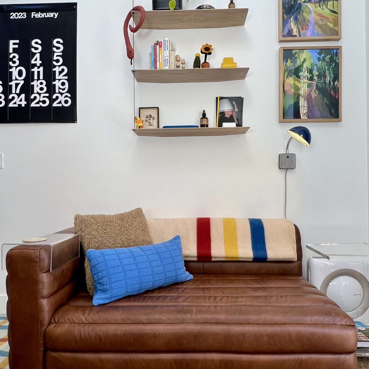 A brown leather sofa with a blue pillow and a striped throw sits against a wall adorned with shelves and framed artwork.