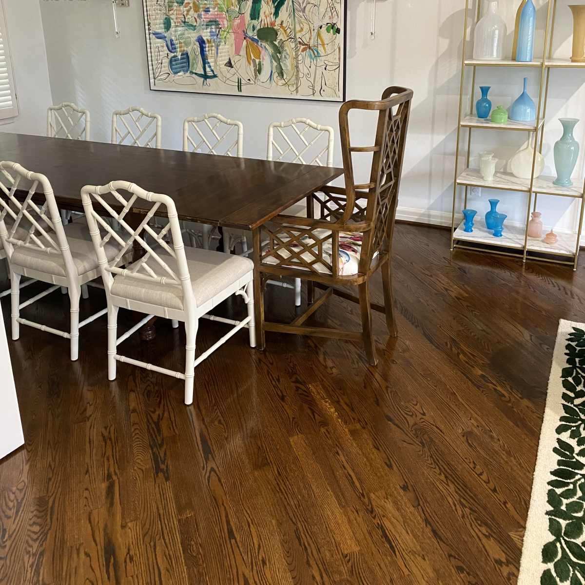 A dining room features a long wooden table surrounded by white chairs, with a large wall art piece and a glass display shelf filled with various decorative items.