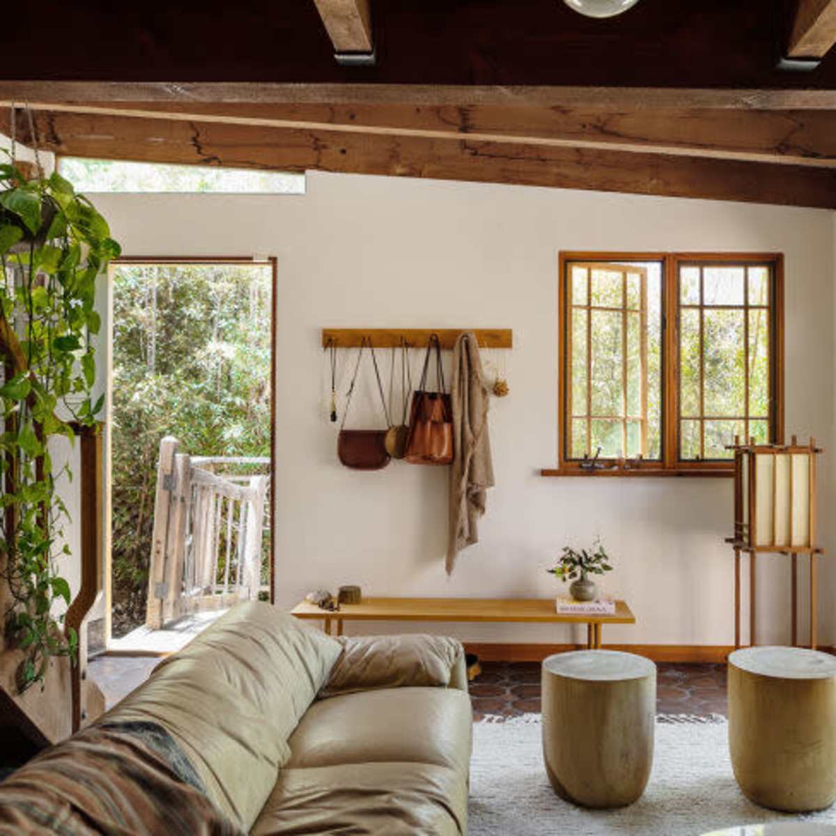 A cozy living room with wooden beams, a large sofa, two round tables, and large windows opening to a green outdoor area.
