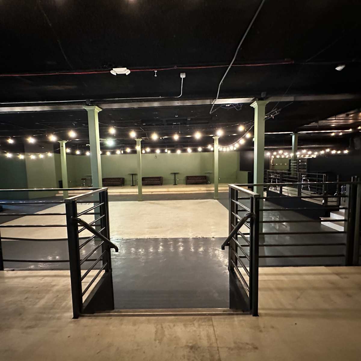 An empty indoor event space with a polished concrete floor, flanked by green walls and overhead string lights.