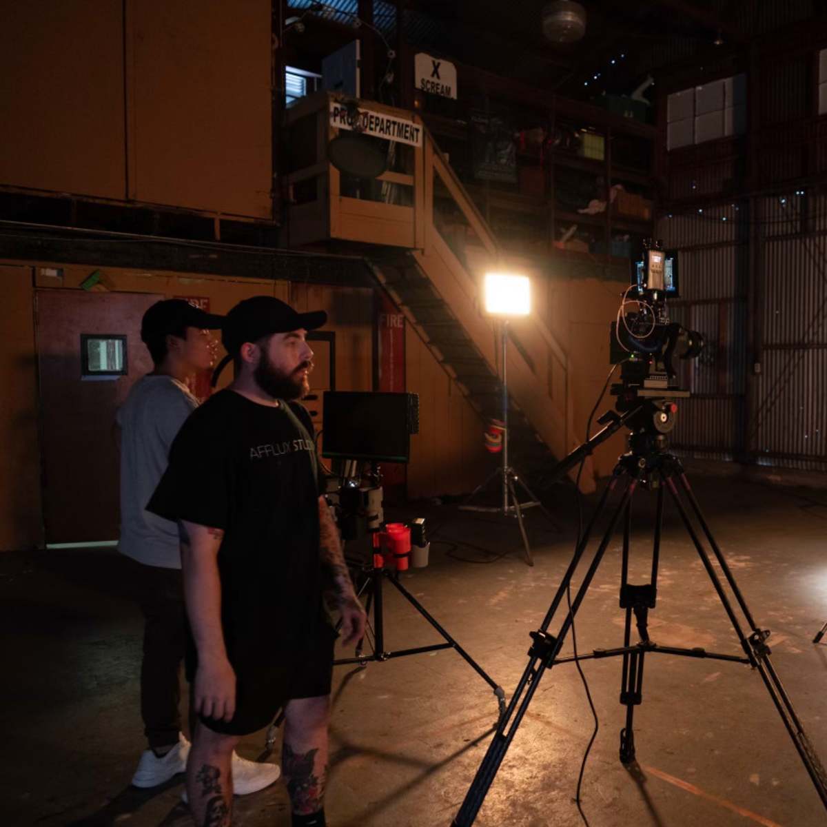 A camera is set up on a tripod in a dimly lit warehouse, with two individuals watching and a third person standing by a lighting setup.