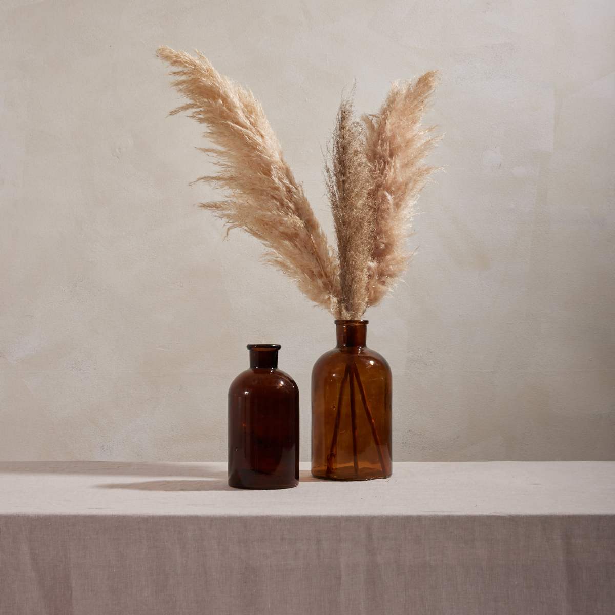 Two brown glass vases with dried pampas grass arranged in one of them sit on a light-colored linen tablecloth against a textured neutral background.