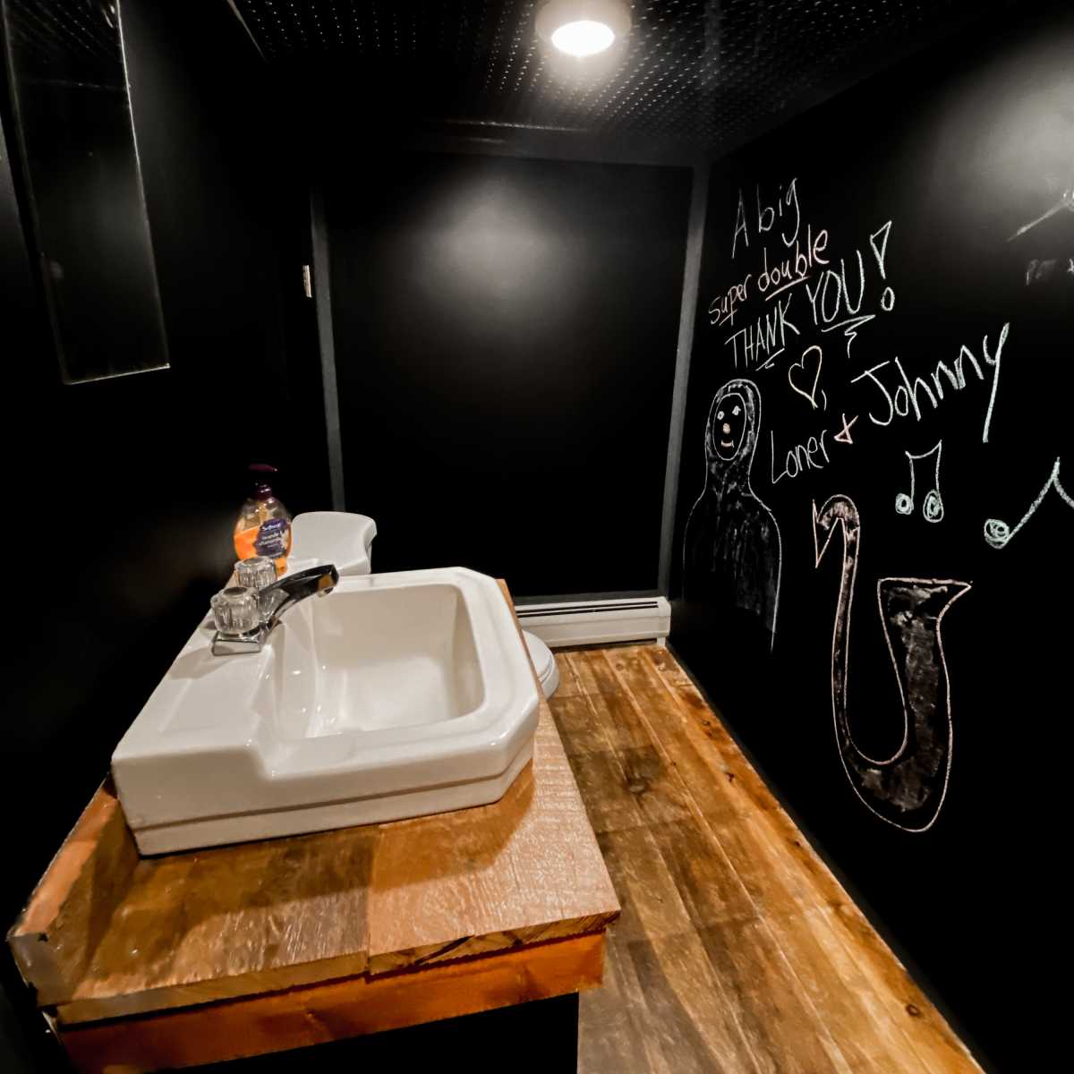 A small, dark bathroom with a white sink on a wooden countertop and a wall covered in chalkboard paint featuring handwritten messages and drawings.