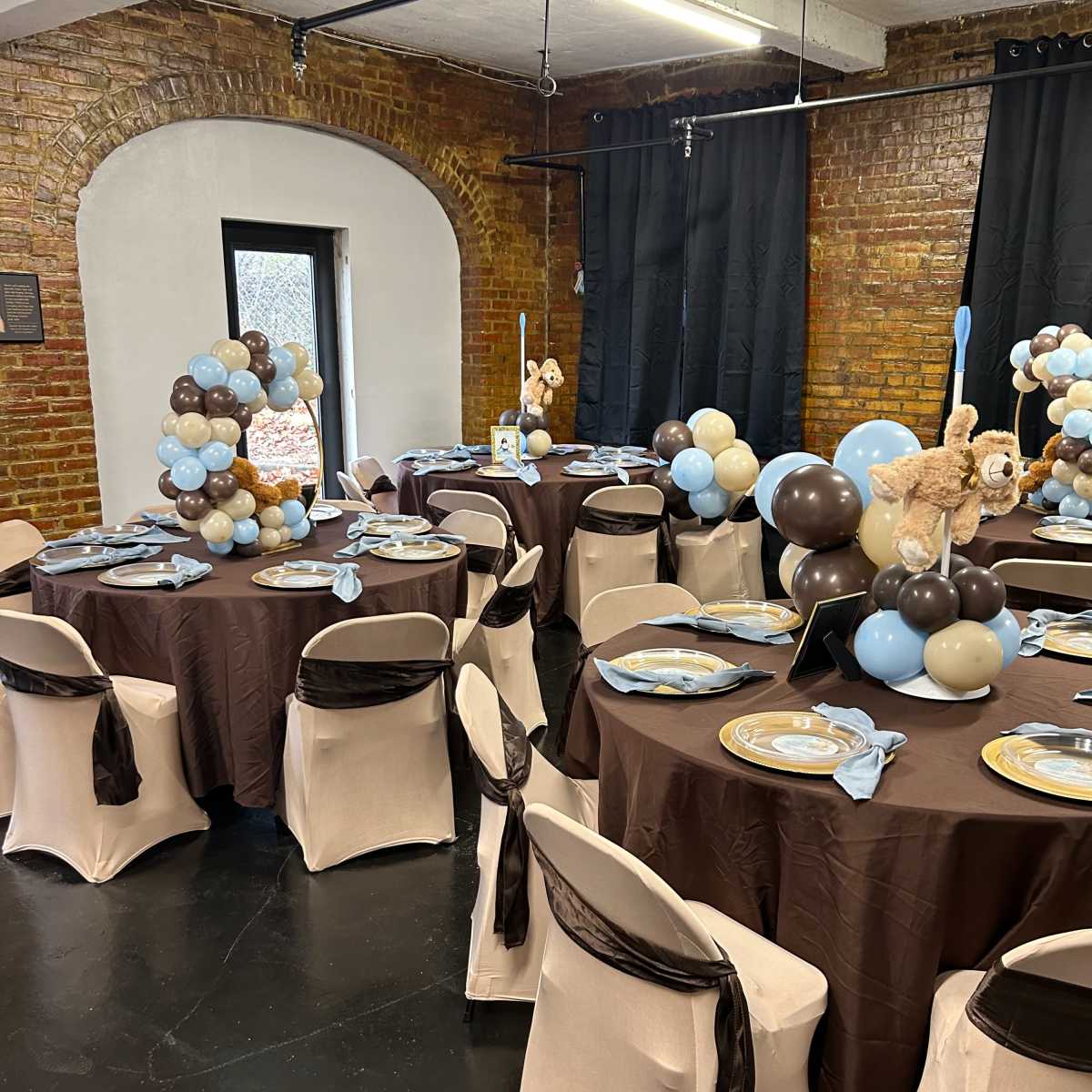 A decorated event space with tables set in brown and cream, featuring centerpieces of balloons and tableware in blue and beige.