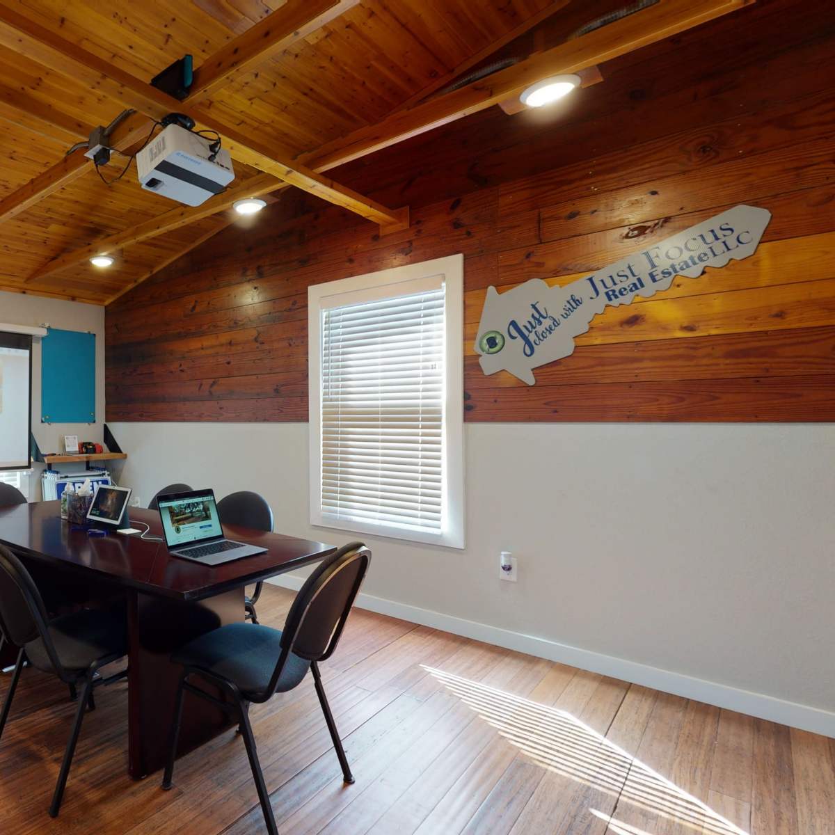 A meeting room with a wooden ceiling, a large projector screen, and a dark table surrounded by chairs, along with a sign that says "Open House."