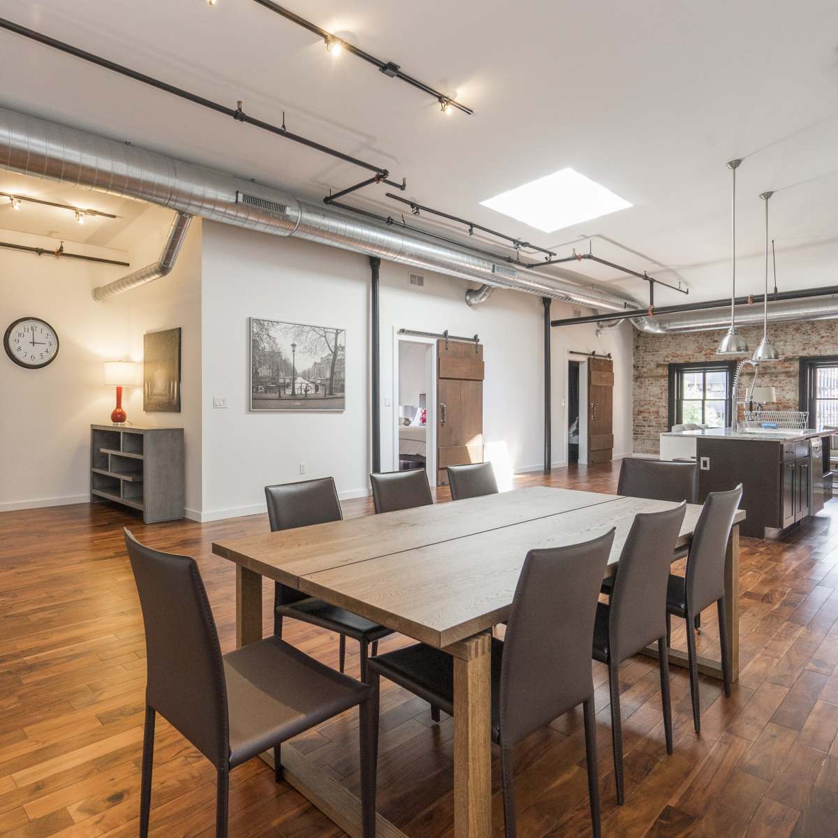 A spacious dining area with a wooden table surrounded by chairs, connected to a modern kitchen and featuring exposed ductwork along the ceiling.