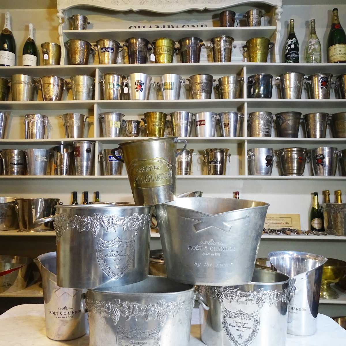 A collection of ornate silver champagne buckets arranged on a marble surface, with a display of various bottles and buckets in the background on shelves.