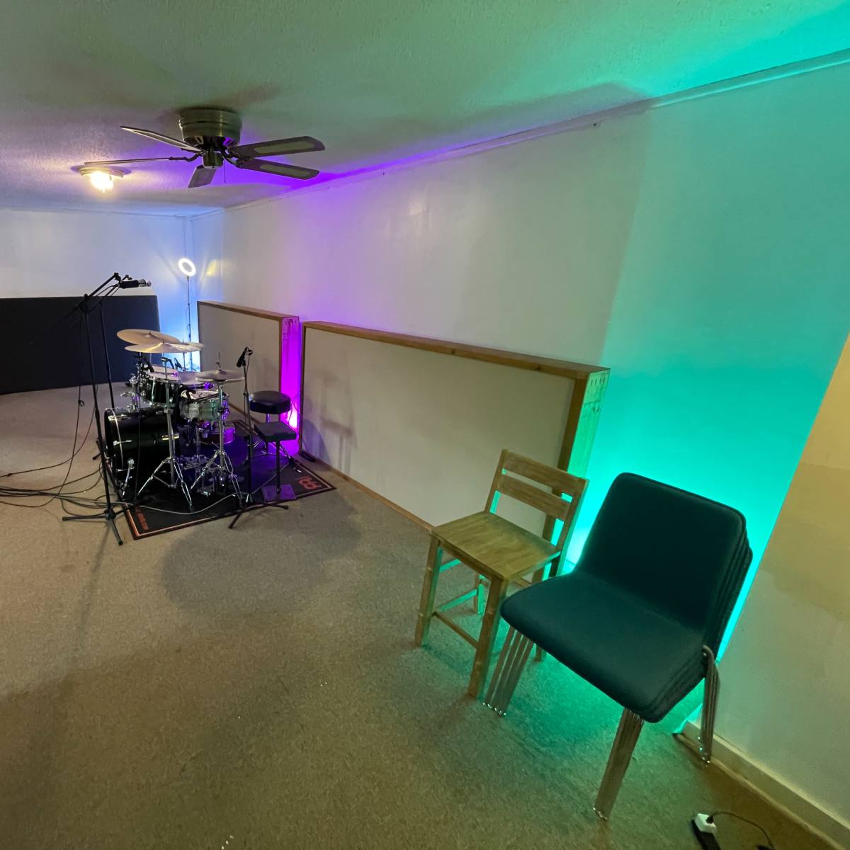 A music rehearsal space with a drum set, microphones, and colorful lighting, alongside two chairs and audio equipment.